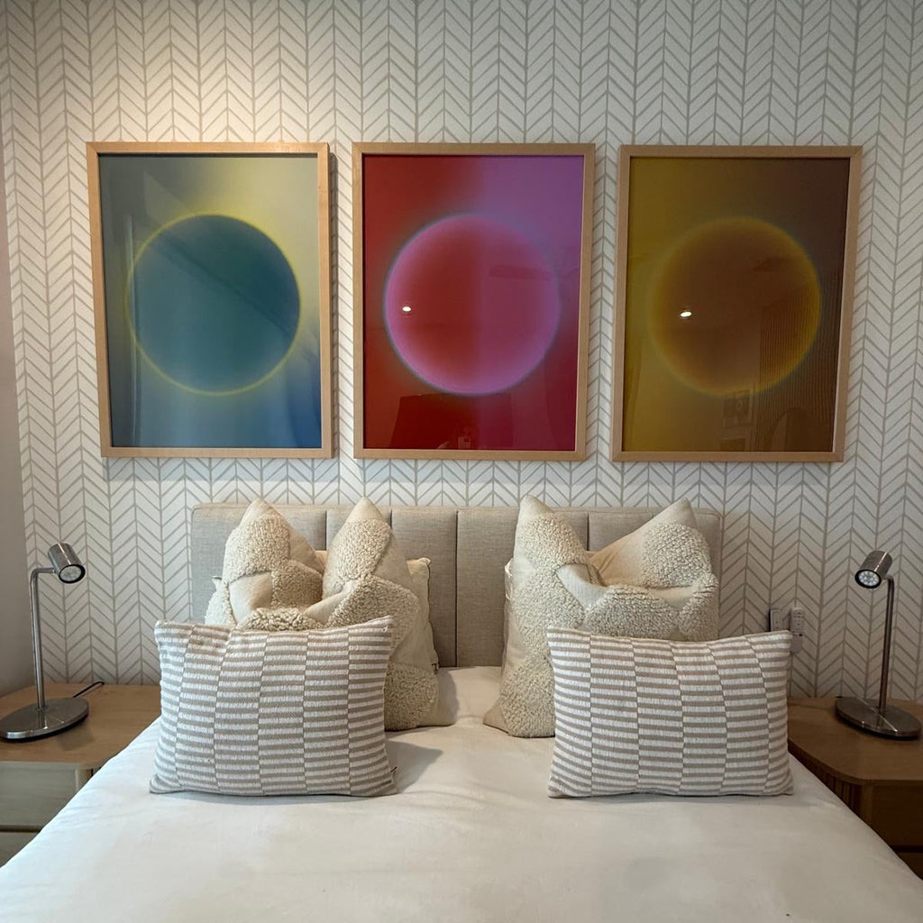A triptych of three Solar series art prints—including Solar N°1 in gold—hanging above a bed in a modern bedroom with a chevron-patterned wallpaper and neutral-toned textured pillows.