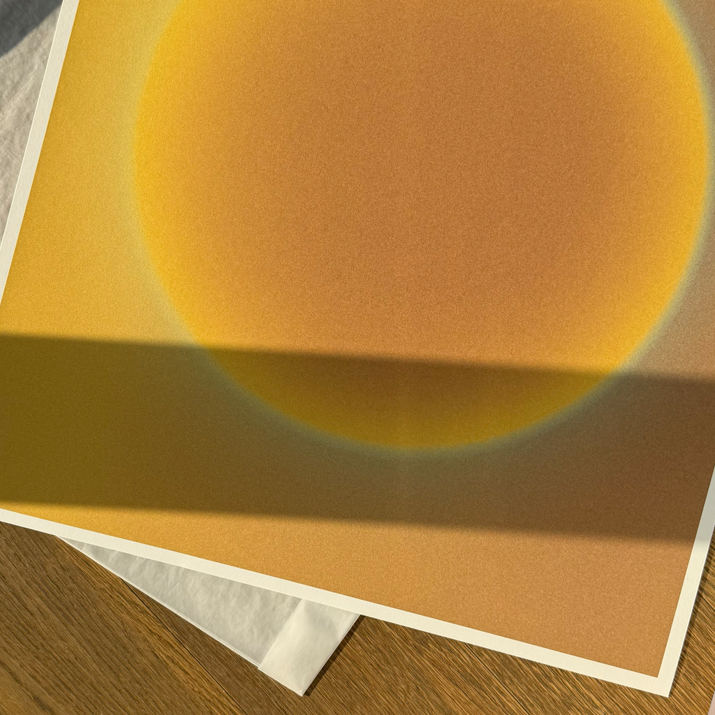 Close-up of the Solar N°1 art print resting on a wooden surface, showing the rich grain of the paper and a glowing golden sun gradient illuminated by soft, natural sunlight and shadows.